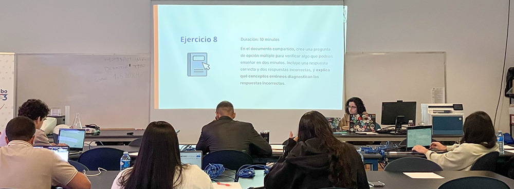 Foto en el aula durante el taller. De espaldas 6 estudiantes sentados trabajando en computadoras. De frente María dando la clase acompañada de una presentación proyectada con el título “Ejercicio 8”
